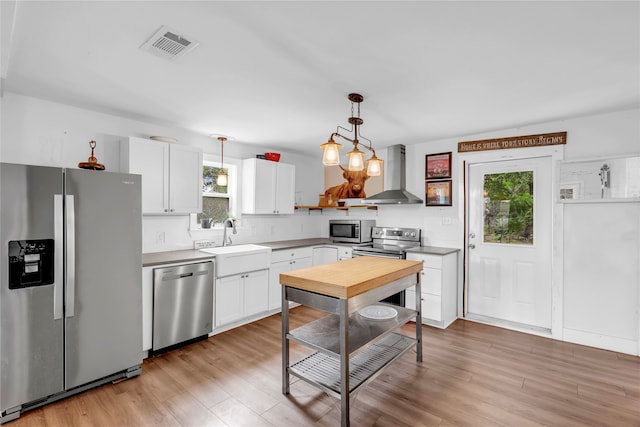 kitchen featuring light wood-type flooring, decorative light fixtures, appliances with stainless steel finishes, white cabinets, and wall chimney range hood