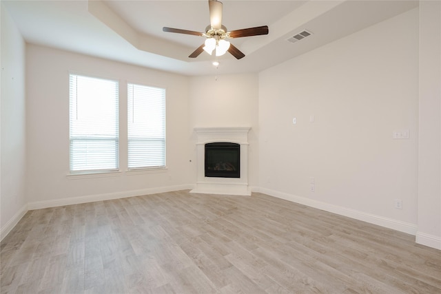 unfurnished living room featuring visible vents, baseboards, a tray ceiling, light wood-style flooring, and a glass covered fireplace