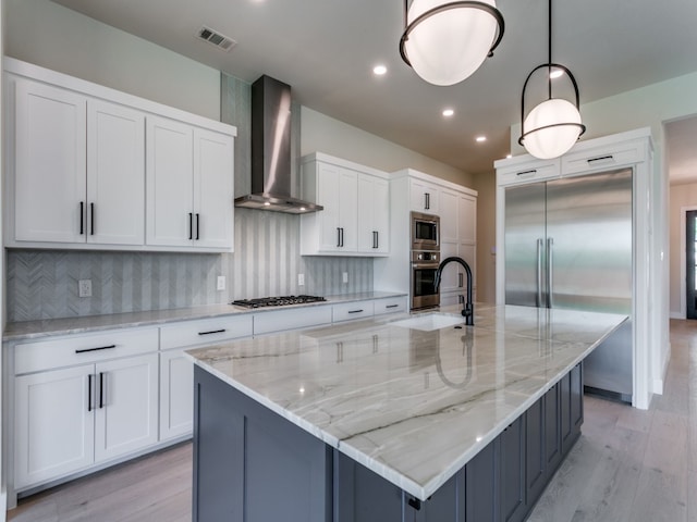 kitchen featuring a large island with sink, wall chimney range hood, built in appliances, decorative light fixtures, and white cabinetry