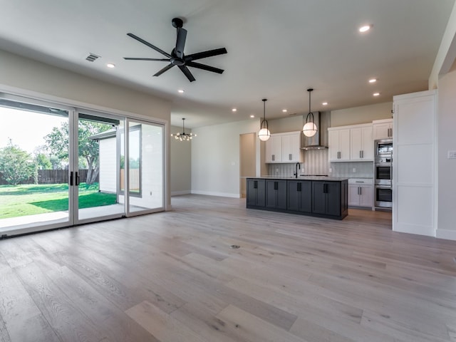 kitchen featuring an island with sink, white cabinetry, light hardwood / wood-style floors, wall chimney exhaust hood, and pendant lighting