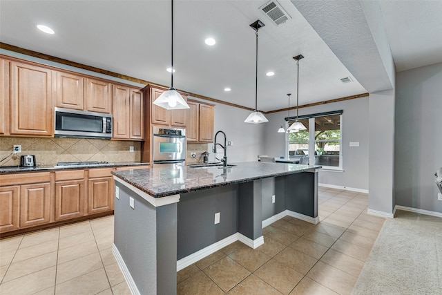 kitchen with dark stone countertops, hanging light fixtures, appliances with stainless steel finishes, sink, and a kitchen island with sink