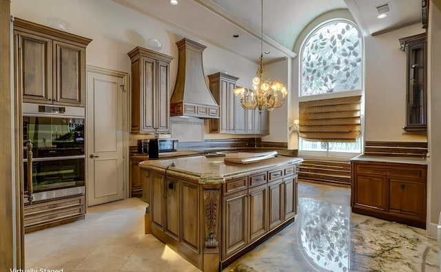 kitchen with light stone countertops, custom exhaust hood, stainless steel double oven, an inviting chandelier, and a center island