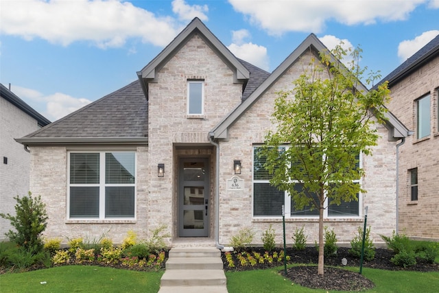 french country style house with brick siding, a shingled roof, and a front yard