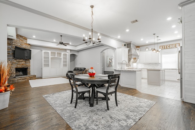 dining area featuring ornamental molding, ceiling fan with notable chandelier, a stone fireplace, and light wood-type flooring