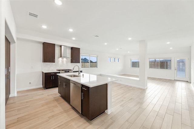 kitchen with wall chimney exhaust hood, sink, a center island with sink, light hardwood / wood-style flooring, and appliances with stainless steel finishes