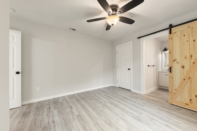 unfurnished bedroom featuring light hardwood / wood-style flooring, ceiling fan, a barn door, and ensuite bathroom