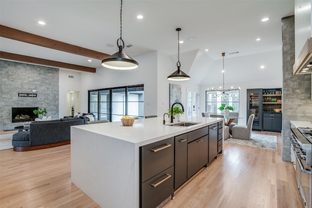 kitchen featuring decorative light fixtures, a stone fireplace, an island with sink, sink, and light hardwood / wood-style flooring