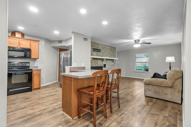 kitchen with light wood-type flooring, a kitchen breakfast bar, black appliances, ceiling fan, and a stone fireplace