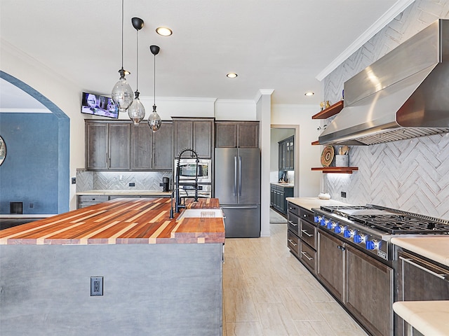 kitchen featuring wood counters, appliances with stainless steel finishes, wall chimney range hood, a center island with sink, and tasteful backsplash