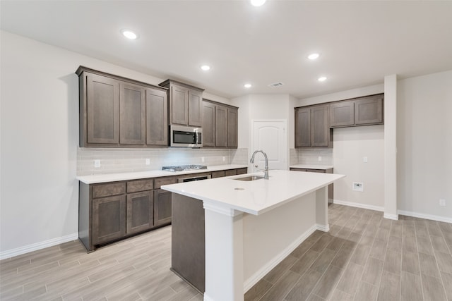 kitchen with dark brown cabinetry, sink, appliances with stainless steel finishes, a kitchen island with sink, and light hardwood / wood-style flooring