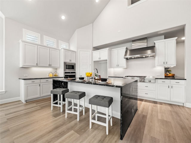 kitchen with stainless steel appliances, wall chimney range hood, white cabinetry, and a kitchen island with sink