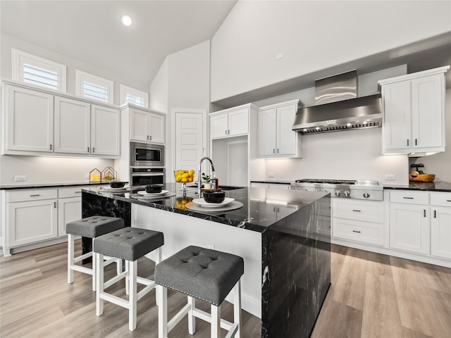 kitchen with a center island with sink, light hardwood / wood-style flooring, appliances with stainless steel finishes, dark stone countertops, and wall chimney range hood