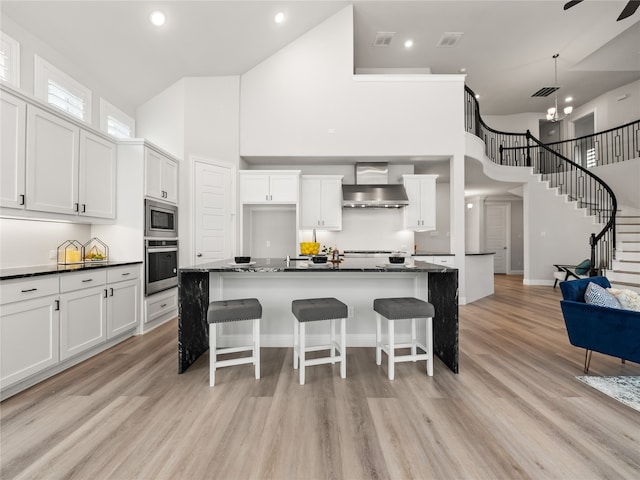 kitchen with appliances with stainless steel finishes, white cabinetry, wall chimney range hood, a center island with sink, and a towering ceiling