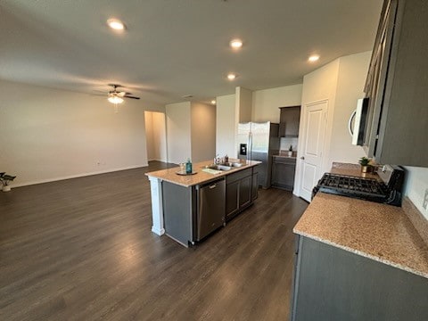 kitchen featuring light stone counters, appliances with stainless steel finishes, dark hardwood / wood-style floors, ceiling fan, and a kitchen island with sink