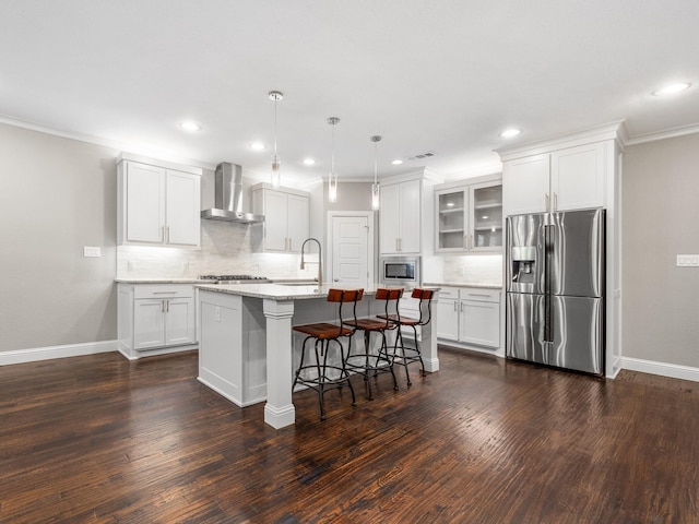 kitchen with light stone countertops, stainless steel appliances, white cabinetry, wall chimney range hood, and an island with sink