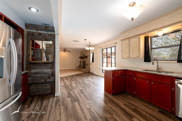 kitchen with sink, dark wood-type flooring, a stone fireplace, ceiling fan with notable chandelier, and appliances with stainless steel finishes