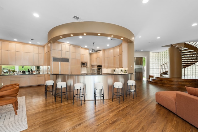 kitchen with a kitchen breakfast bar, light brown cabinetry, hanging light fixtures, appliances with stainless steel finishes, and hardwood / wood-style flooring