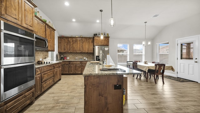 kitchen featuring appliances with stainless steel finishes, light stone countertops, lofted ceiling, a center island with sink, and tasteful backsplash