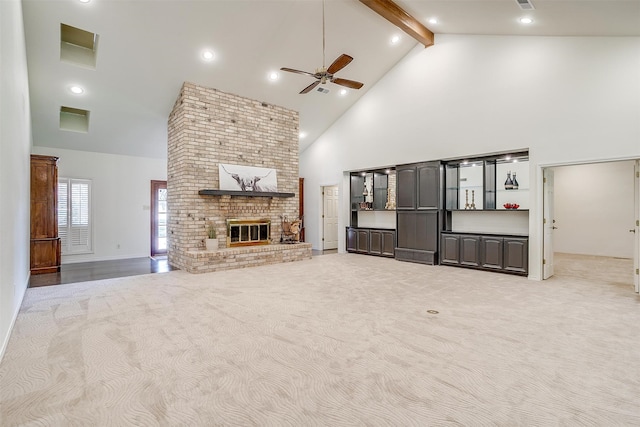 unfurnished living room featuring beamed ceiling, high vaulted ceiling, a brick fireplace, light colored carpet, and ceiling fan