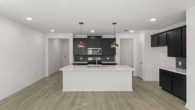 kitchen featuring appliances with stainless steel finishes, pendant lighting, decorative backsplash, a center island with sink, and light wood-type flooring