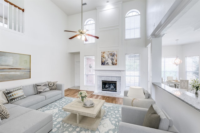 living room with a high ceiling, a tiled fireplace, ceiling fan with notable chandelier, and light hardwood / wood-style floors