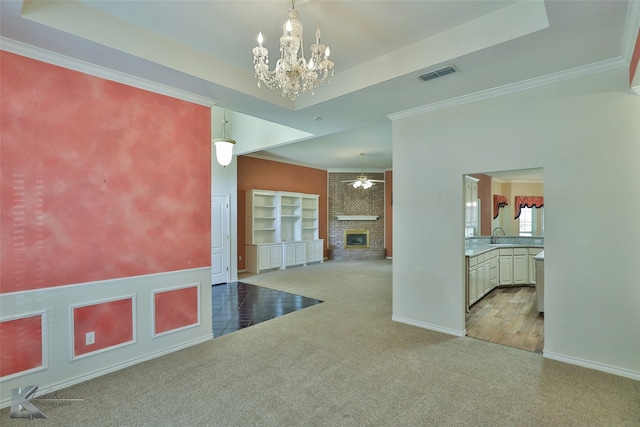 unfurnished room featuring ceiling fan with notable chandelier, ornamental molding, a brick fireplace, carpet flooring, and a tray ceiling