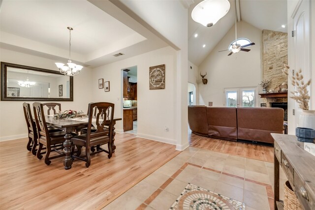 dining space with a stone fireplace, light hardwood / wood-style flooring, ceiling fan with notable chandelier, and high vaulted ceiling