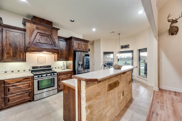 kitchen featuring backsplash, stainless steel appliances, an island with sink, custom range hood, and dark brown cabinetry