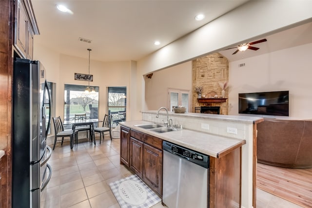 kitchen with light wood-type flooring, ceiling fan with notable chandelier, stainless steel appliances, sink, and a stone fireplace