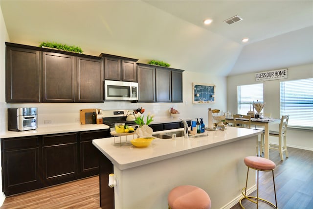 kitchen featuring vaulted ceiling, stainless steel appliances, light hardwood / wood-style floors, an island with sink, and a kitchen breakfast bar