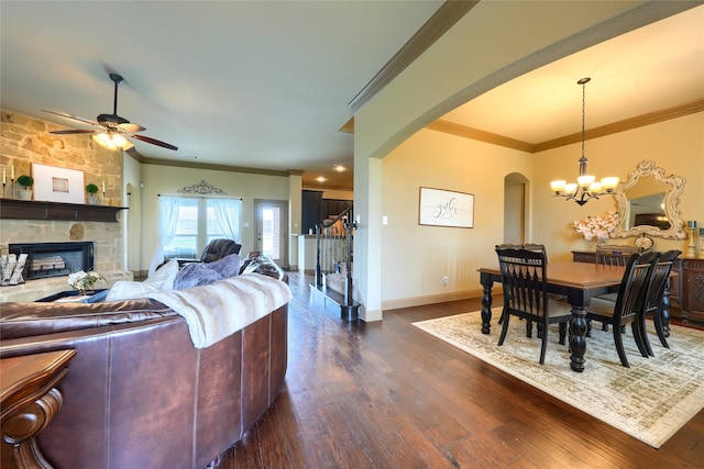 living room featuring dark wood-type flooring, ornamental molding, ceiling fan with notable chandelier, and a stone fireplace