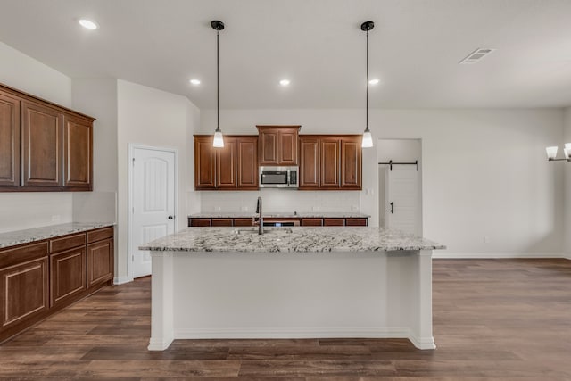 kitchen featuring hanging light fixtures, a barn door, light stone counters, dark hardwood / wood-style floors, and a center island with sink