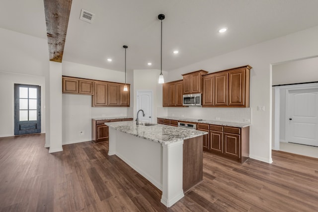 kitchen with decorative light fixtures, appliances with stainless steel finishes, light stone counters, dark hardwood / wood-style floors, and an island with sink