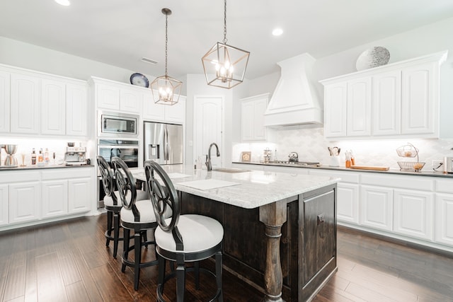 kitchen featuring custom range hood, appliances with stainless steel finishes, light stone counters, dark hardwood / wood-style floors, and a kitchen island with sink