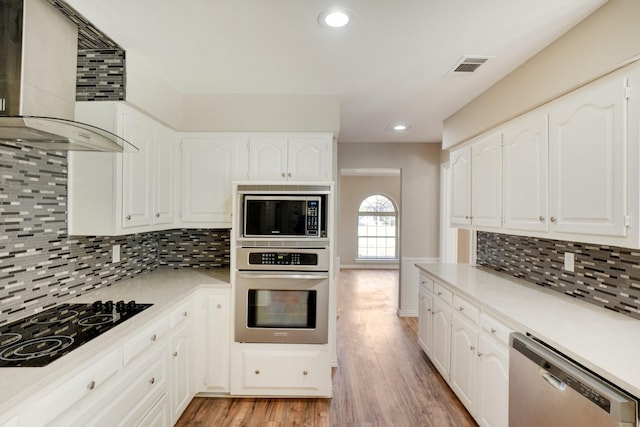 kitchen featuring wall chimney range hood, appliances with stainless steel finishes, white cabinets, decorative backsplash, and light wood-type flooring