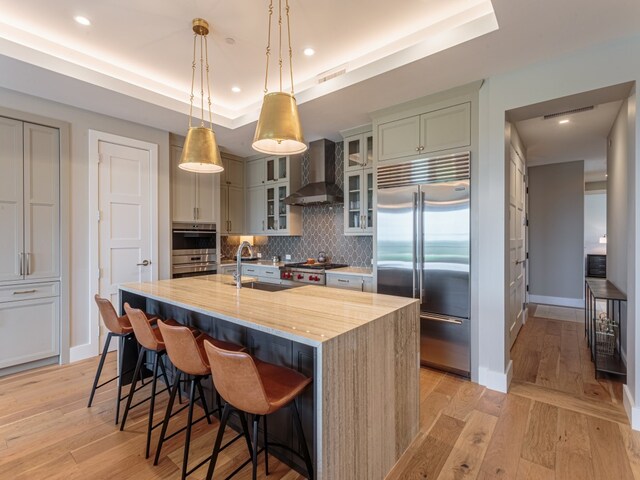 kitchen with a kitchen island with sink, sink, wall chimney exhaust hood, appliances with stainless steel finishes, and light hardwood / wood-style floors