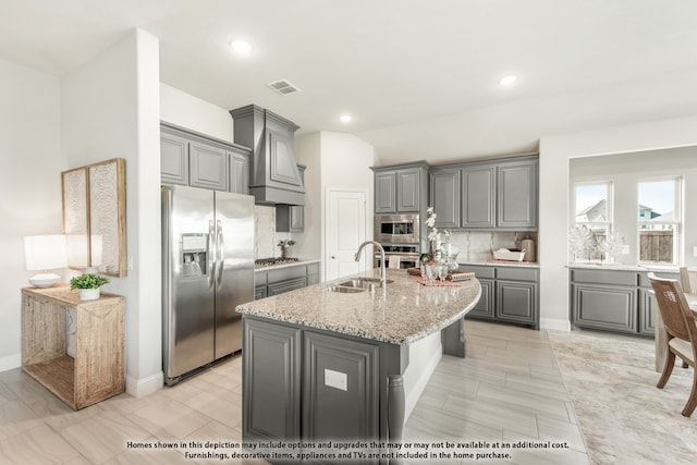 kitchen with custom exhaust hood, a center island with sink, sink, gray cabinetry, and stainless steel appliances