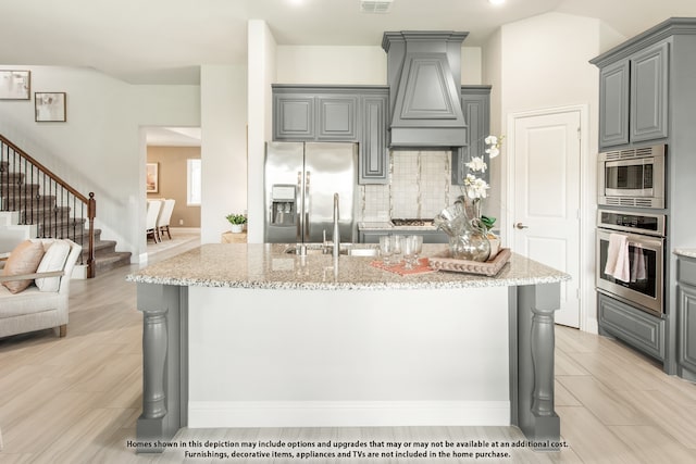 kitchen with an island with sink, appliances with stainless steel finishes, light stone countertops, gray cabinets, and premium range hood