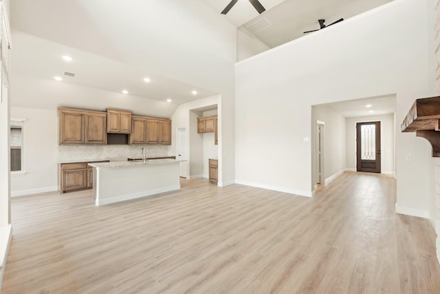 kitchen with light wood-type flooring, a kitchen island with sink, a towering ceiling, ceiling fan, and decorative backsplash