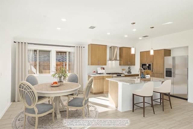 kitchen featuring sink, wall chimney range hood, hanging light fixtures, stainless steel appliances, and a center island with sink