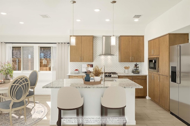 kitchen with stainless steel appliances, hanging light fixtures, wall chimney range hood, and a center island with sink