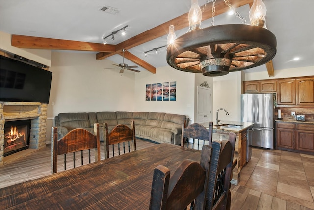 dining space with rail lighting, ceiling fan, sink, a fireplace, and vaulted ceiling with beams