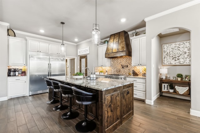 kitchen with appliances with stainless steel finishes, dark wood-type flooring, an island with sink, light stone countertops, and custom exhaust hood