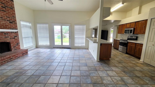 kitchen with a brick fireplace, stainless steel appliances, kitchen peninsula, ceiling fan, and a towering ceiling