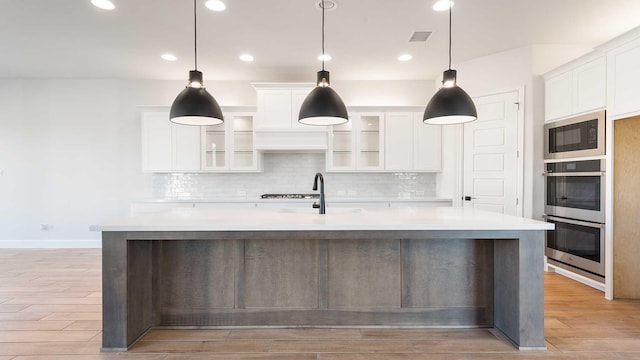 kitchen featuring white cabinets, stainless steel double oven, decorative light fixtures, and a kitchen island with sink