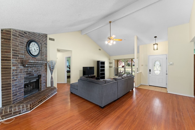 living room with a brick fireplace, beamed ceiling, wood-type flooring, ceiling fan, and a textured ceiling