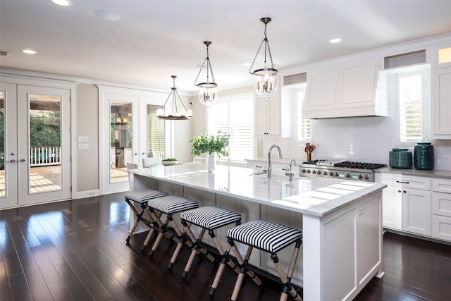 kitchen with premium range hood, a kitchen island with sink, sink, decorative light fixtures, and white cabinetry