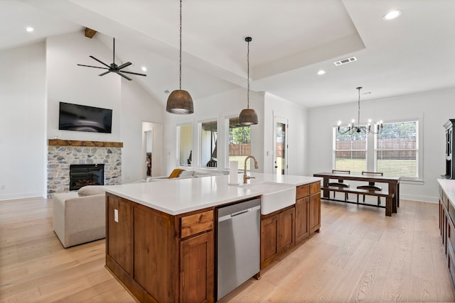 kitchen featuring ceiling fan with notable chandelier, dishwasher, an island with sink, a stone fireplace, and light hardwood / wood-style floors