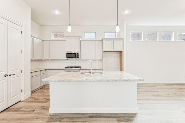 kitchen featuring appliances with stainless steel finishes, light stone counters, a kitchen island with sink, sink, and hanging light fixtures