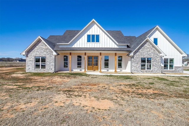 modern farmhouse with covered porch, brick siding, french doors, roof with shingles, and board and batten siding